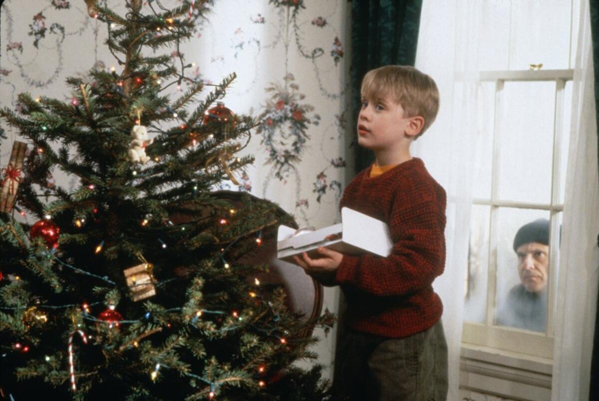 Macaulay Culkin stands in front of a Christmas tree as Joe Pesci looks through a window from the outside in "Home Alone."
