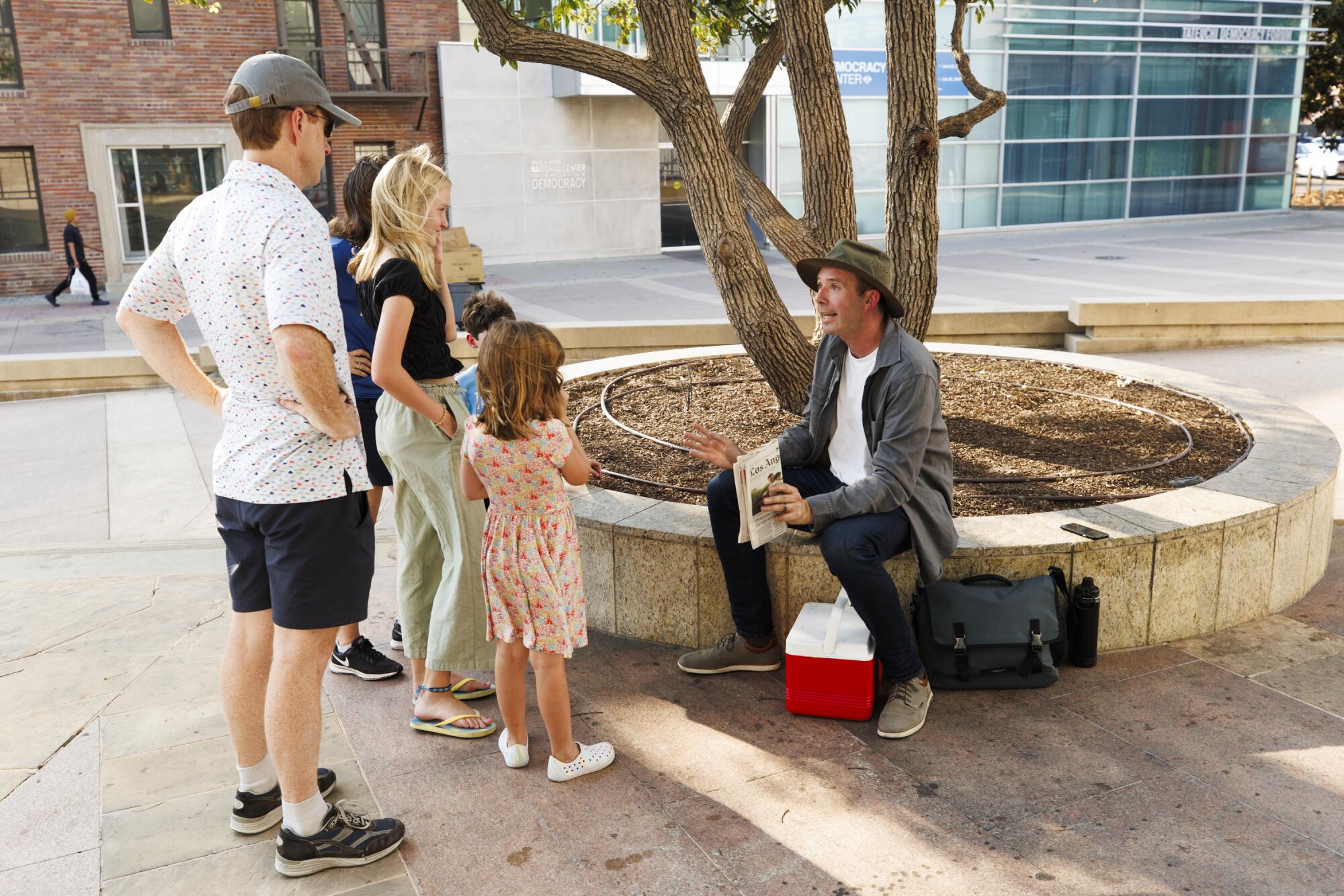 A man sitting on a low wall around a tree talks to a family in front of him.