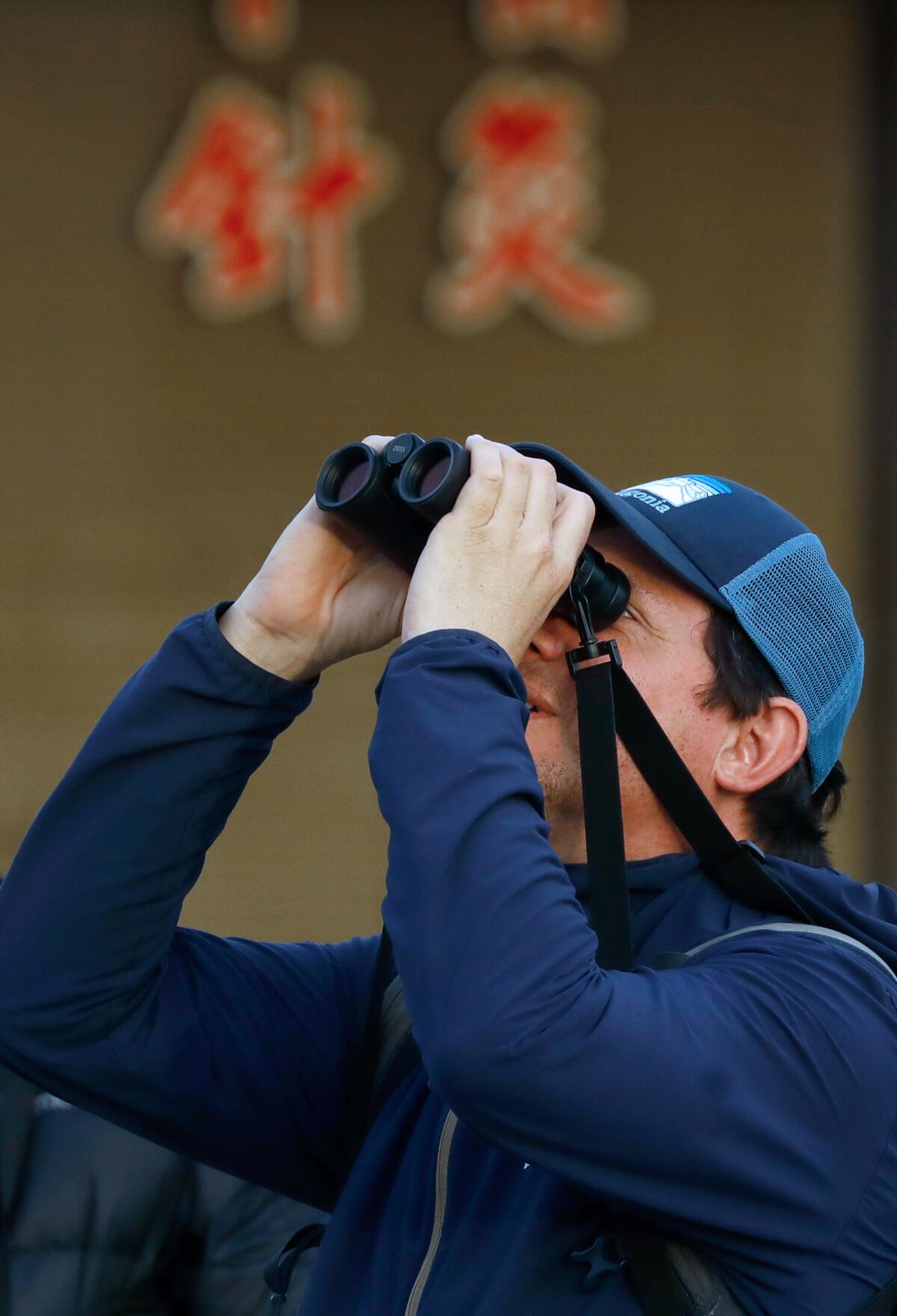 A man wearing a blue jacket and blue baseball cap, looking up through binoculars.