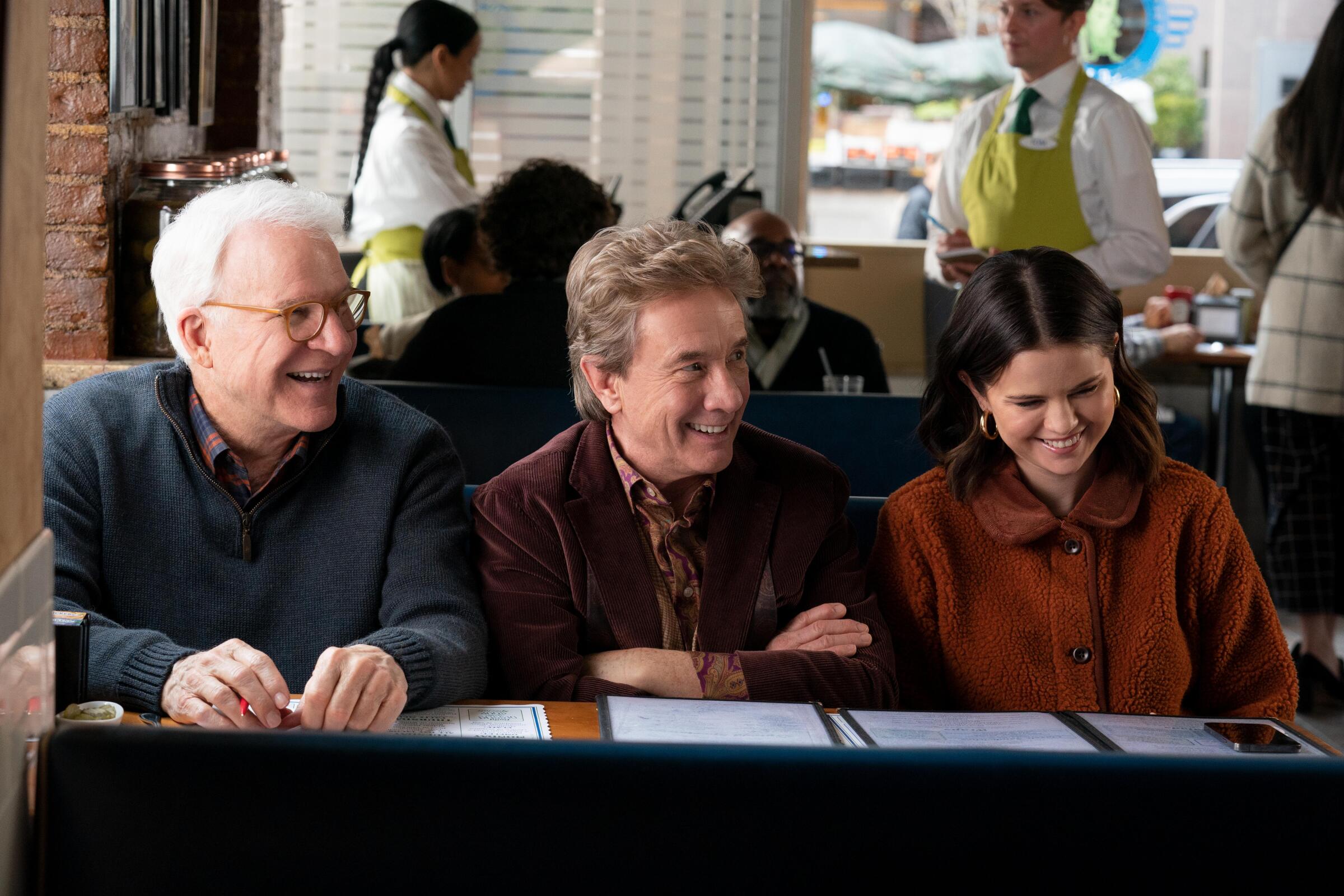 Two older men and a younger woman sit at a diner booth smiling in a scene from "Only Murders in the Building."