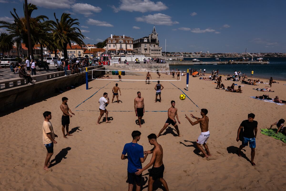 People in swim trunks bat a volleyball near a net. Others sunbathe. In the background are buildings and palm trees.
