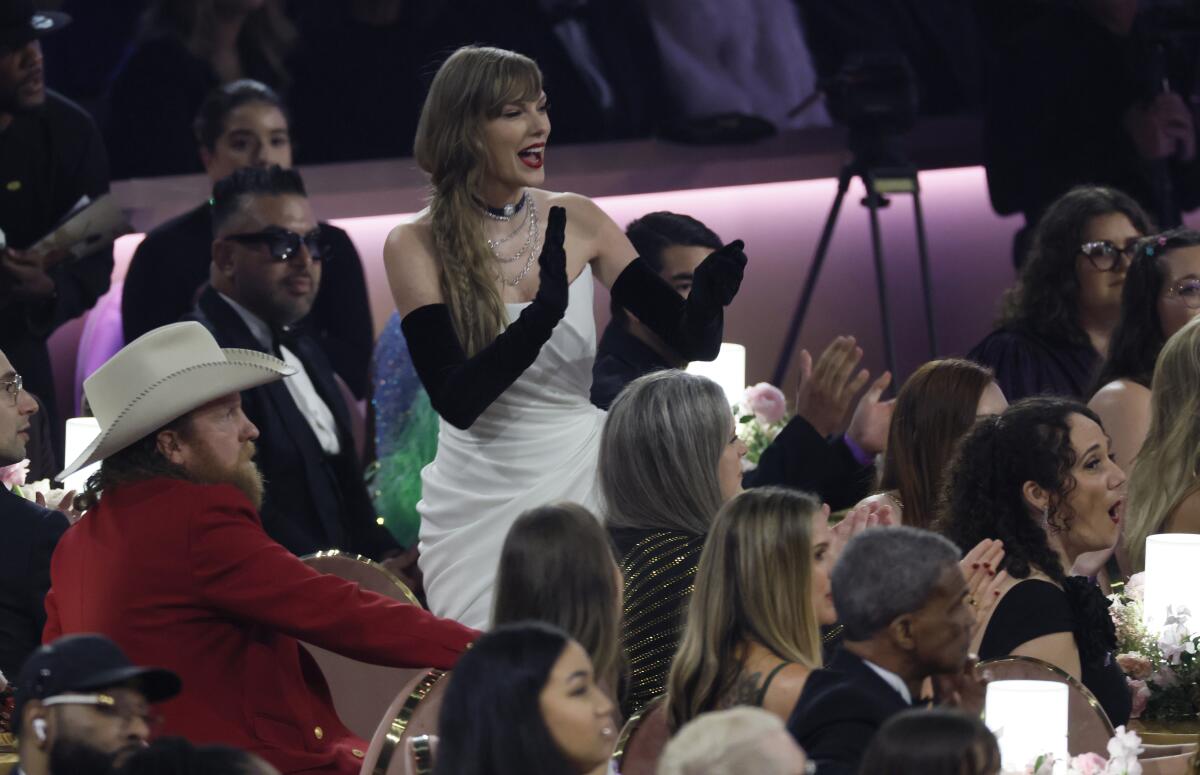 Taylor Swift in opera-length black gloves and strapless white gown stands among people in formal wear seated at tables.