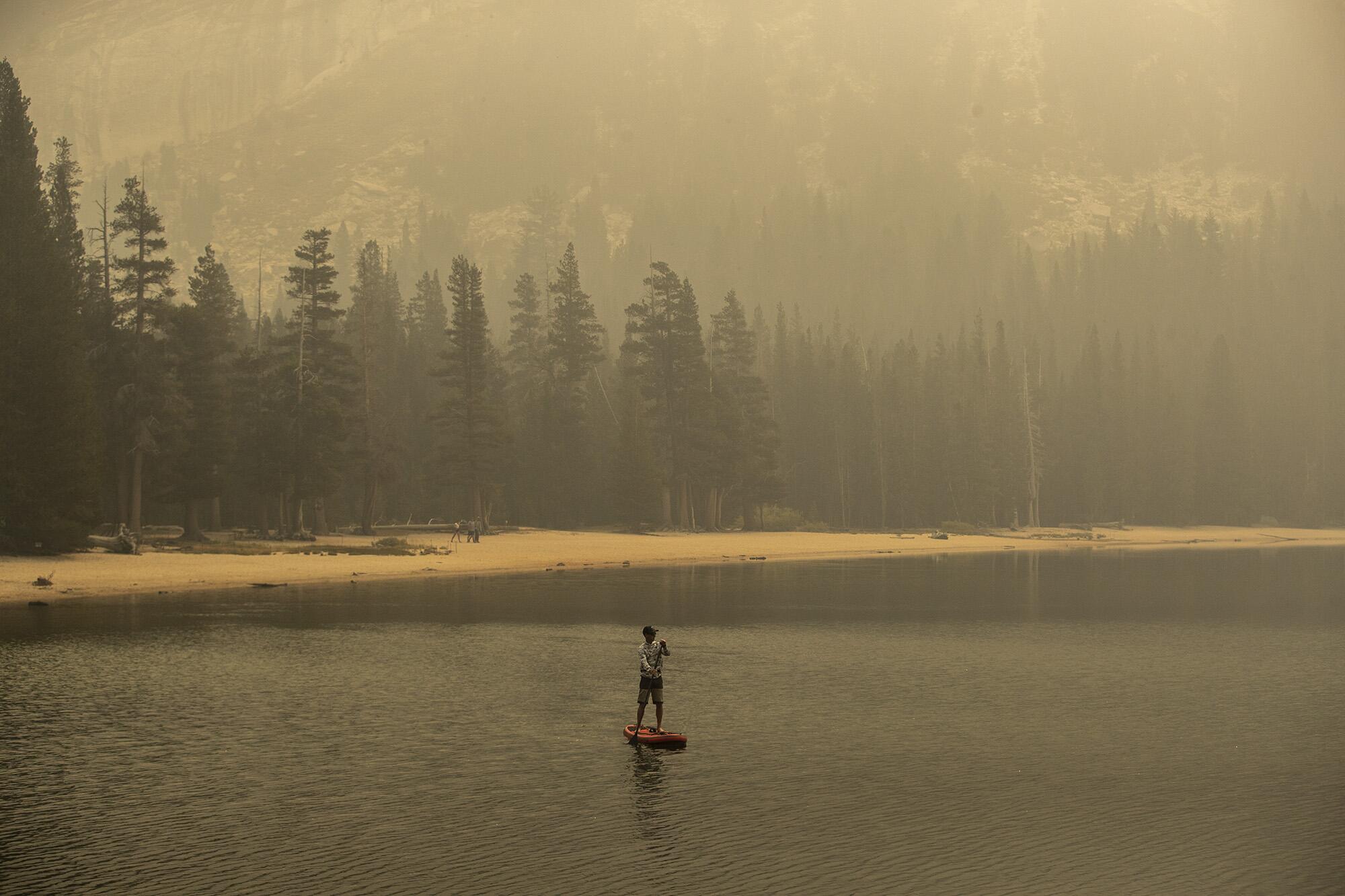 A lone figure paddleboards on a smoke-covered lake.