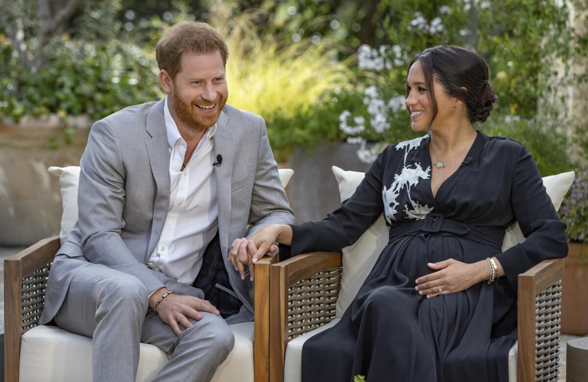 Prince Harry in a casual suit and Meghan, Duchess of Sussex, in a black dress, hold hands  