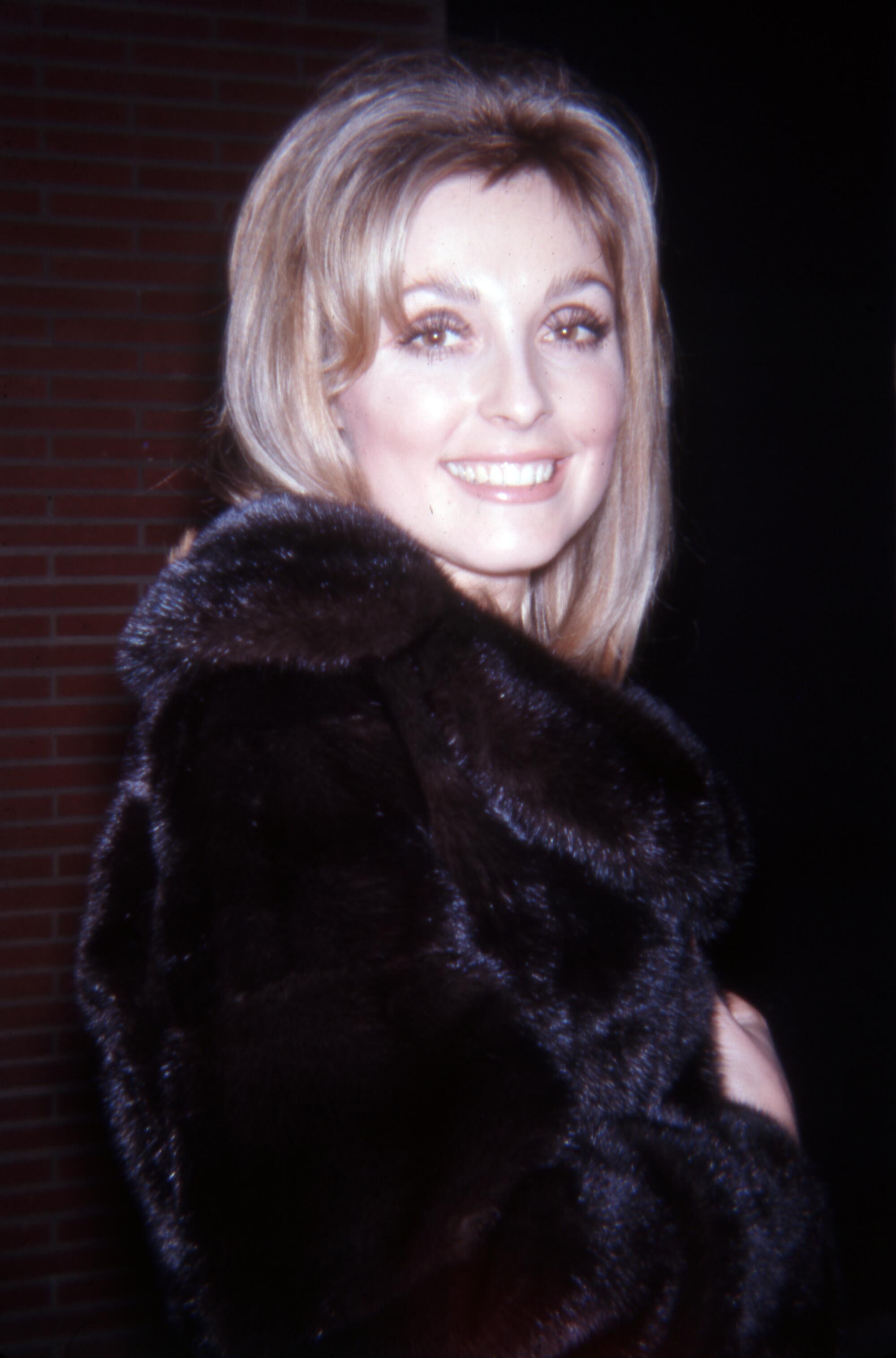 A woman with light brown hair in a dark fur coat smiles toward the camera.
