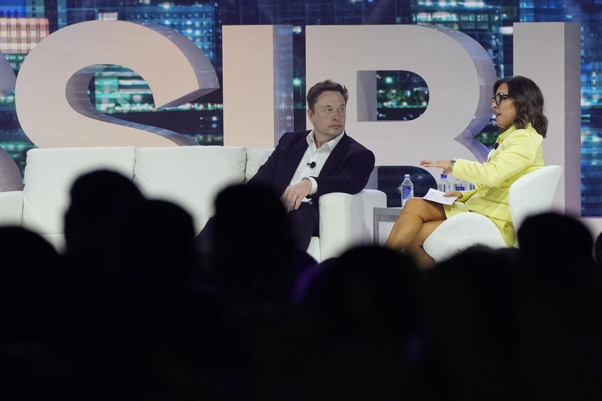 Both seated on a stage in front of an audience, Elon Musk speaks with Linda Yaccarino.