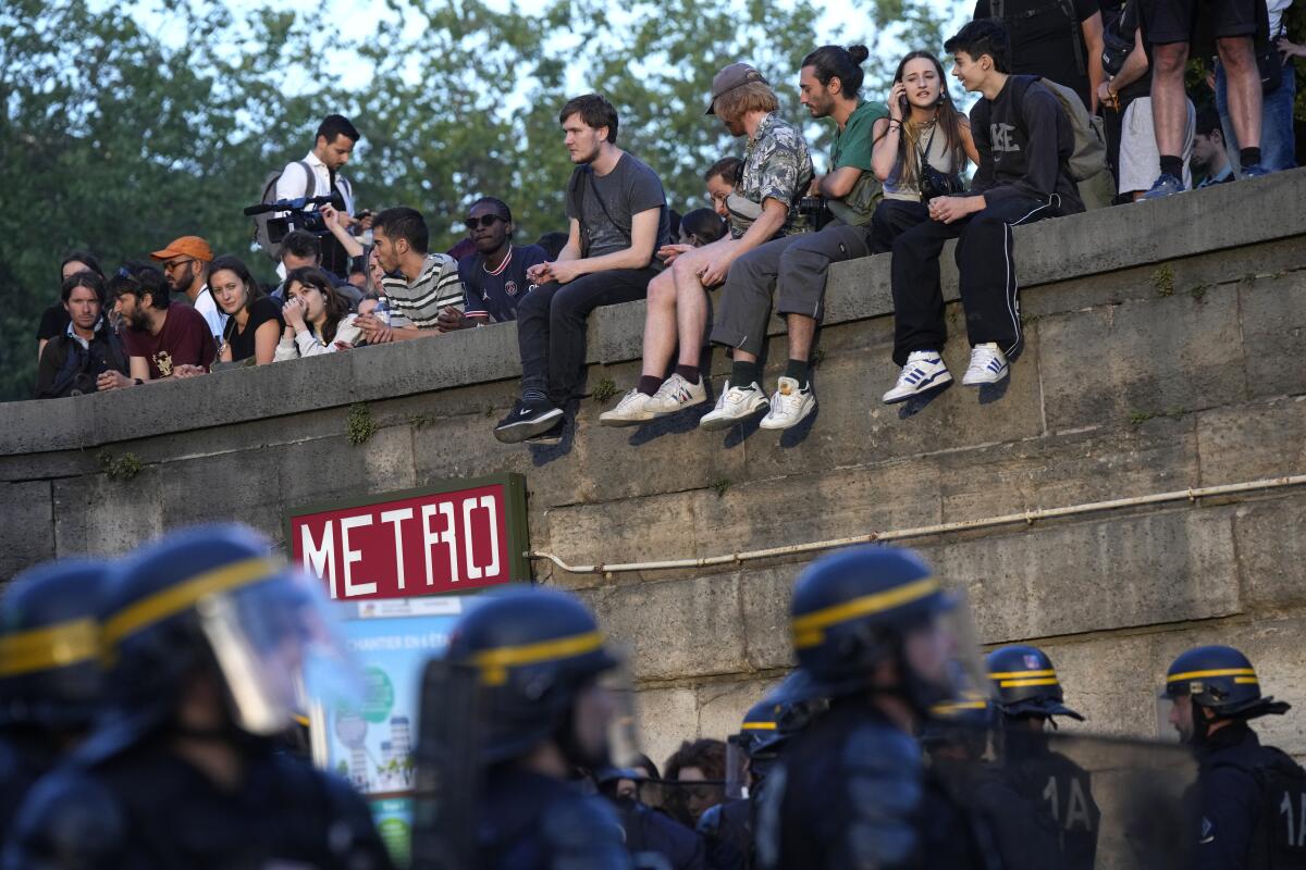Police patrol as young sit or stand nearby during a protest in Paris.