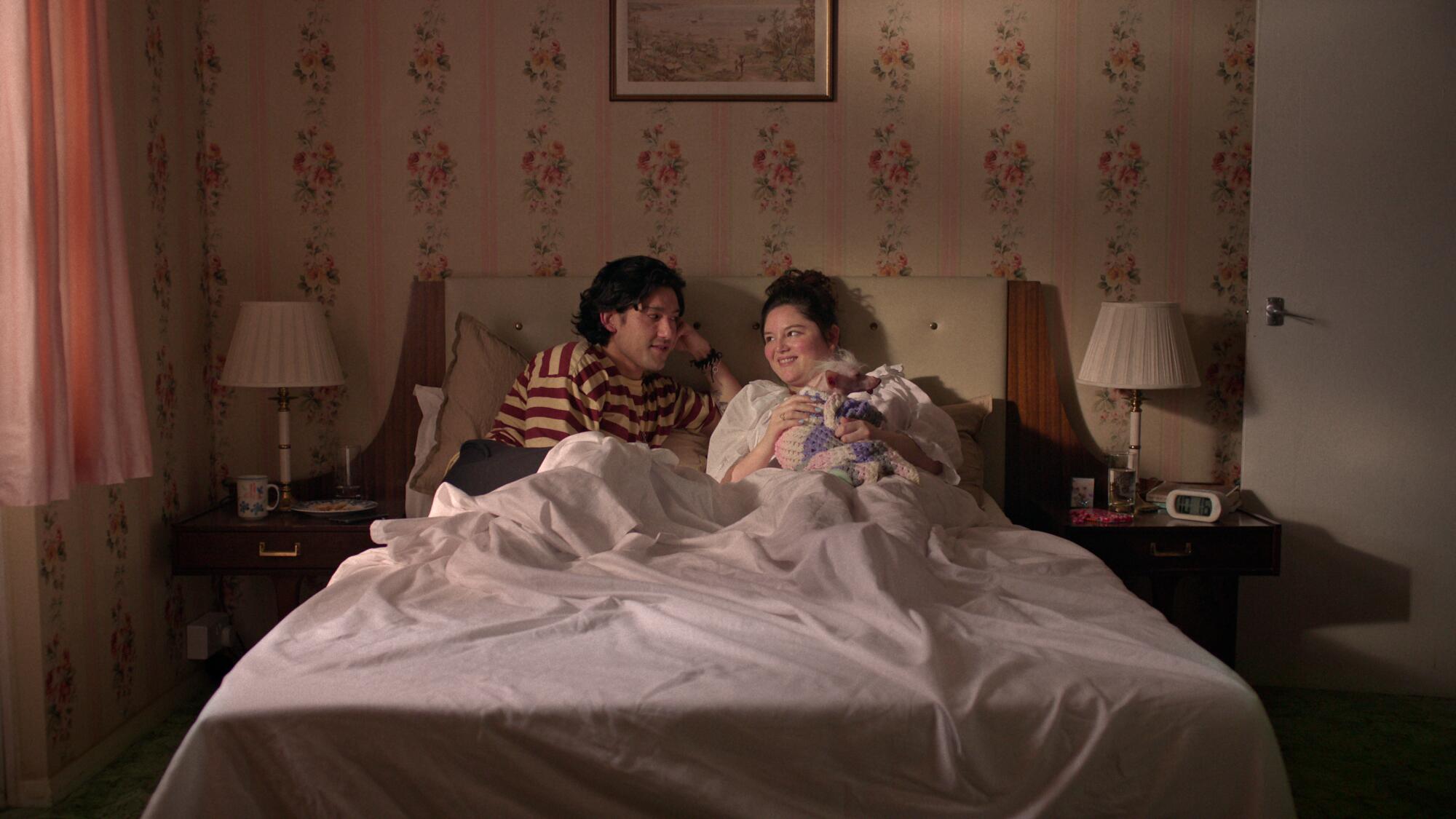 A man in a striped shirt sits in bed next to a woman holding a small dog