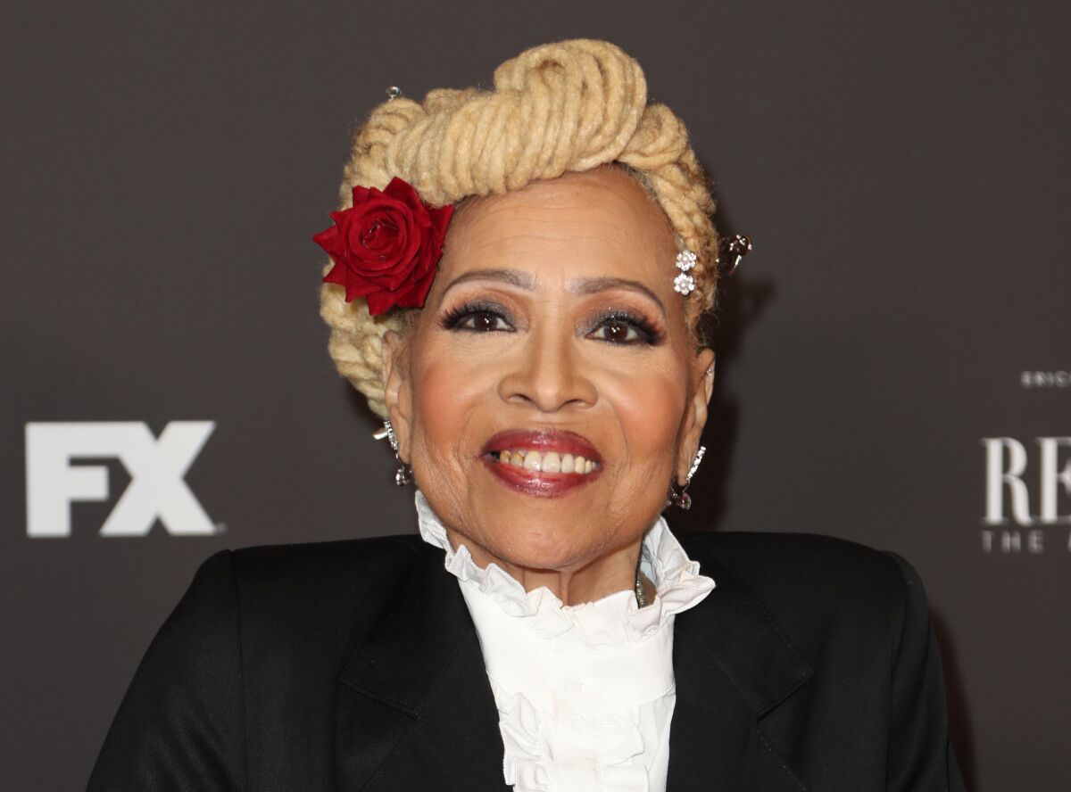 A woman with blond hair wrapped into a bun with a red rose in it posing in a black suit and ruffled white shirt.