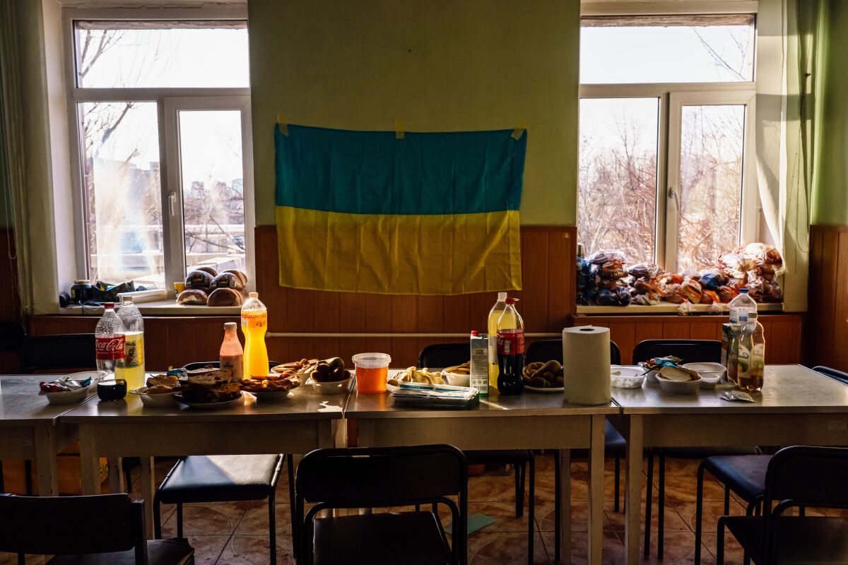 A Ukrainian flag hangs on a wall over a table with various food items — in an empty room illuminated by afternoon sun.