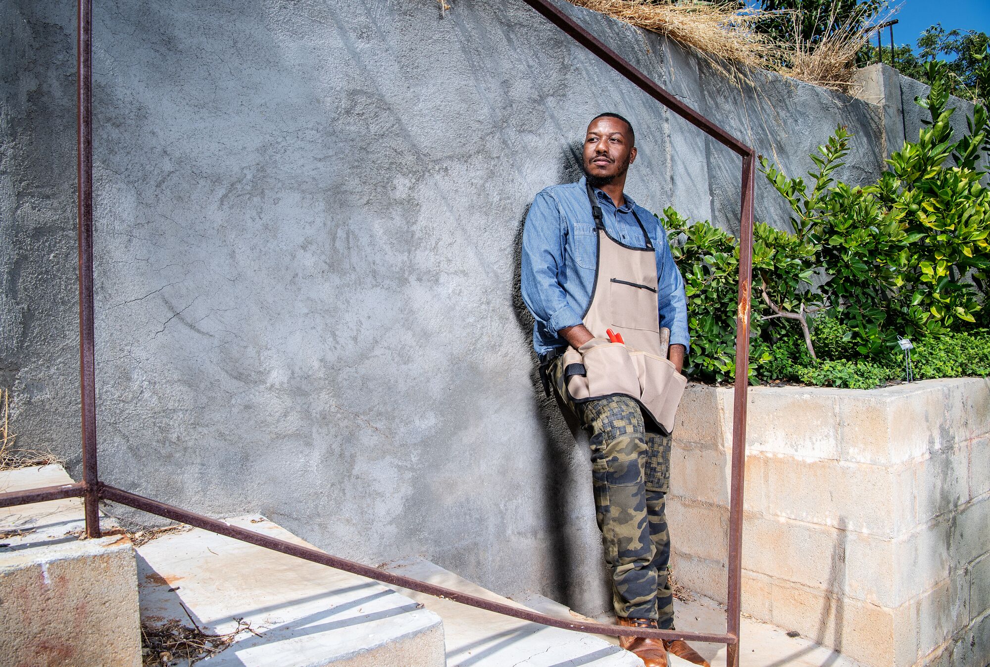 A man wearing camouflage pants and a handyman apron leans against a concrete wall in his garden.
