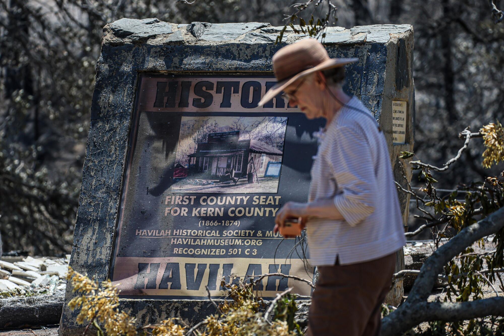 A Havilah resident wearing a sun hat passes a charred sign for the Havilah museum. 