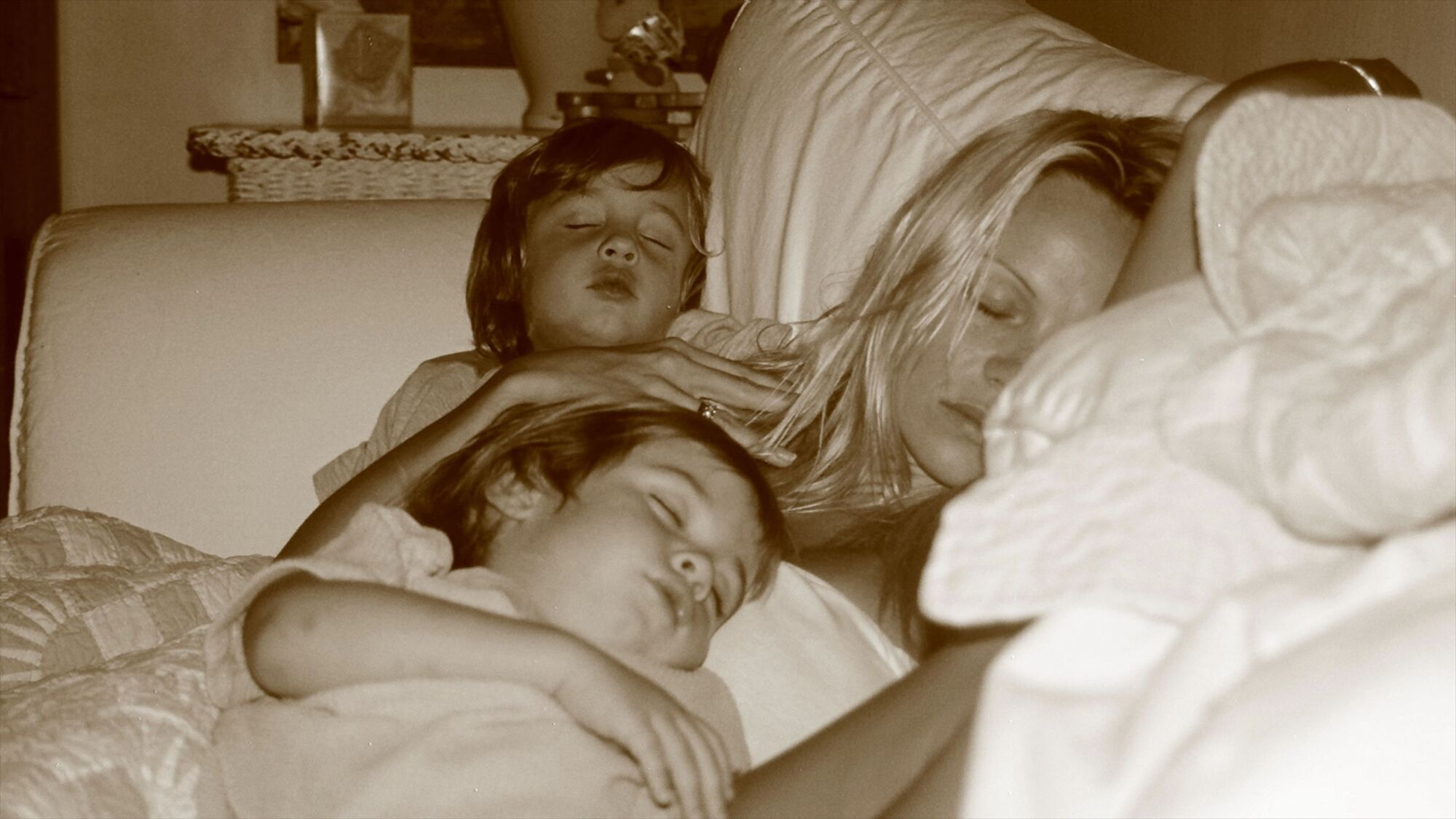 A blond woman sleeps on a sofa with her two sons snuggled around her.