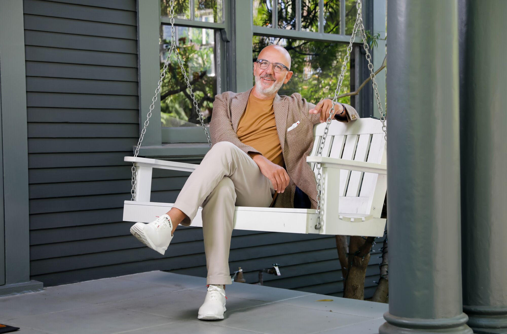 A man sits, legs crossed, on a white porch swing.