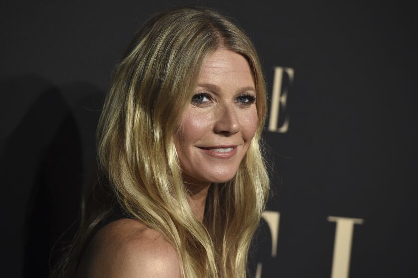 Woman, Gwyneth Paltrow looks over her shoulder with blonde hair down along both sides of her face, giving slight smile