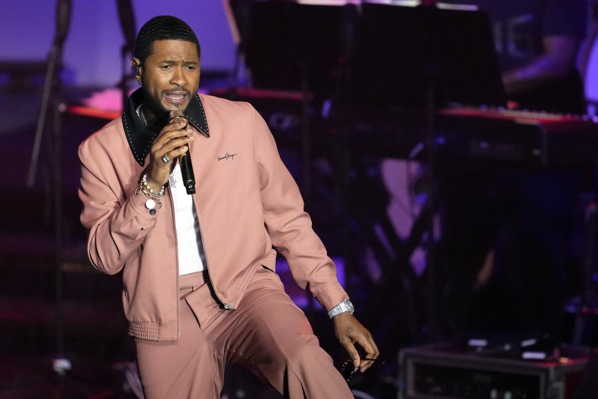 Usher sings into a microphone while performing in a dusty pink jacket and pants