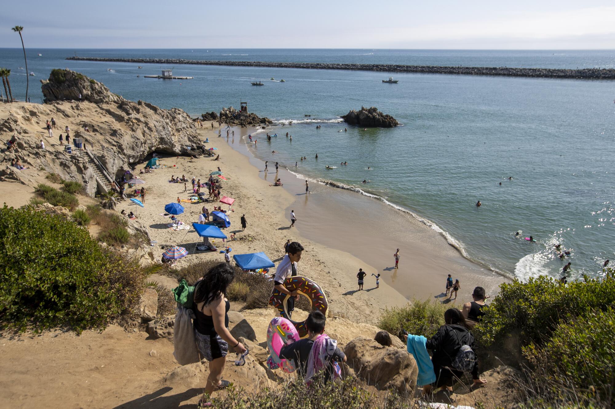 People carrying inflatable floaties climb down a steep, sandy hill toward the beach.