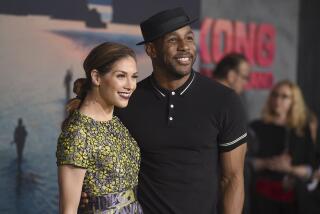 A woman with long brown hair in a yellow dress posing with a man in a black hat and black shirt, both smiling