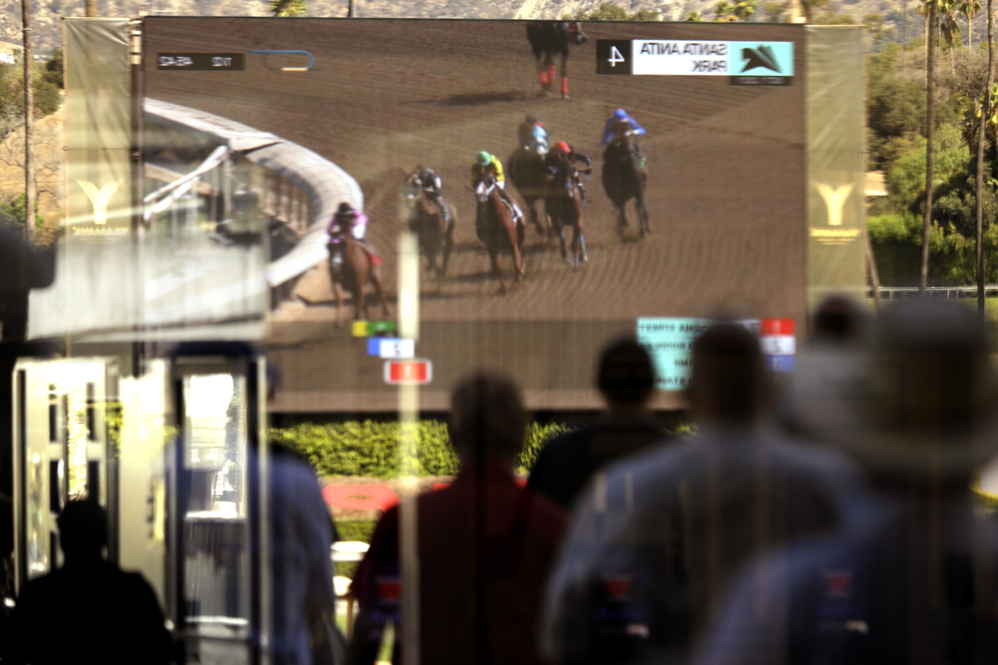 Visitors, in a reflection, watch the fourth race on the big screen on opening day at Santa Anita Park in Arcadia.