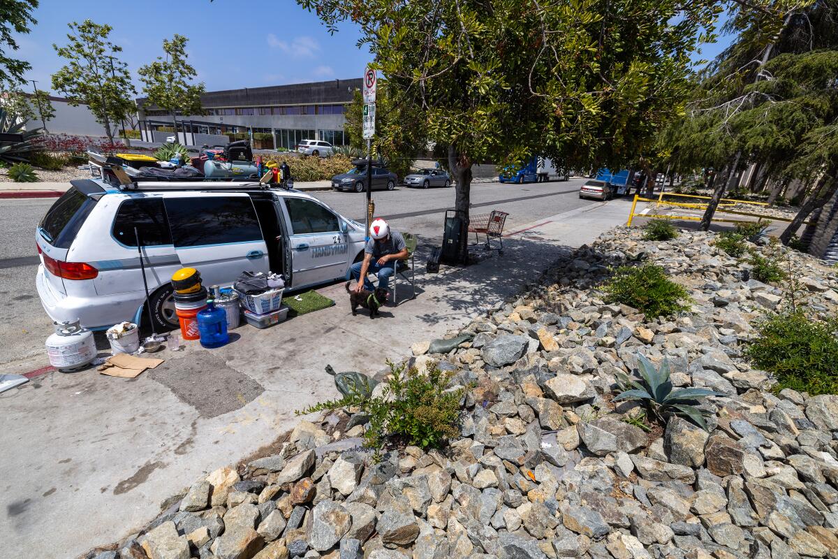 A man pets his dog next to his broken-down van, with belongings placed on the sidewalk