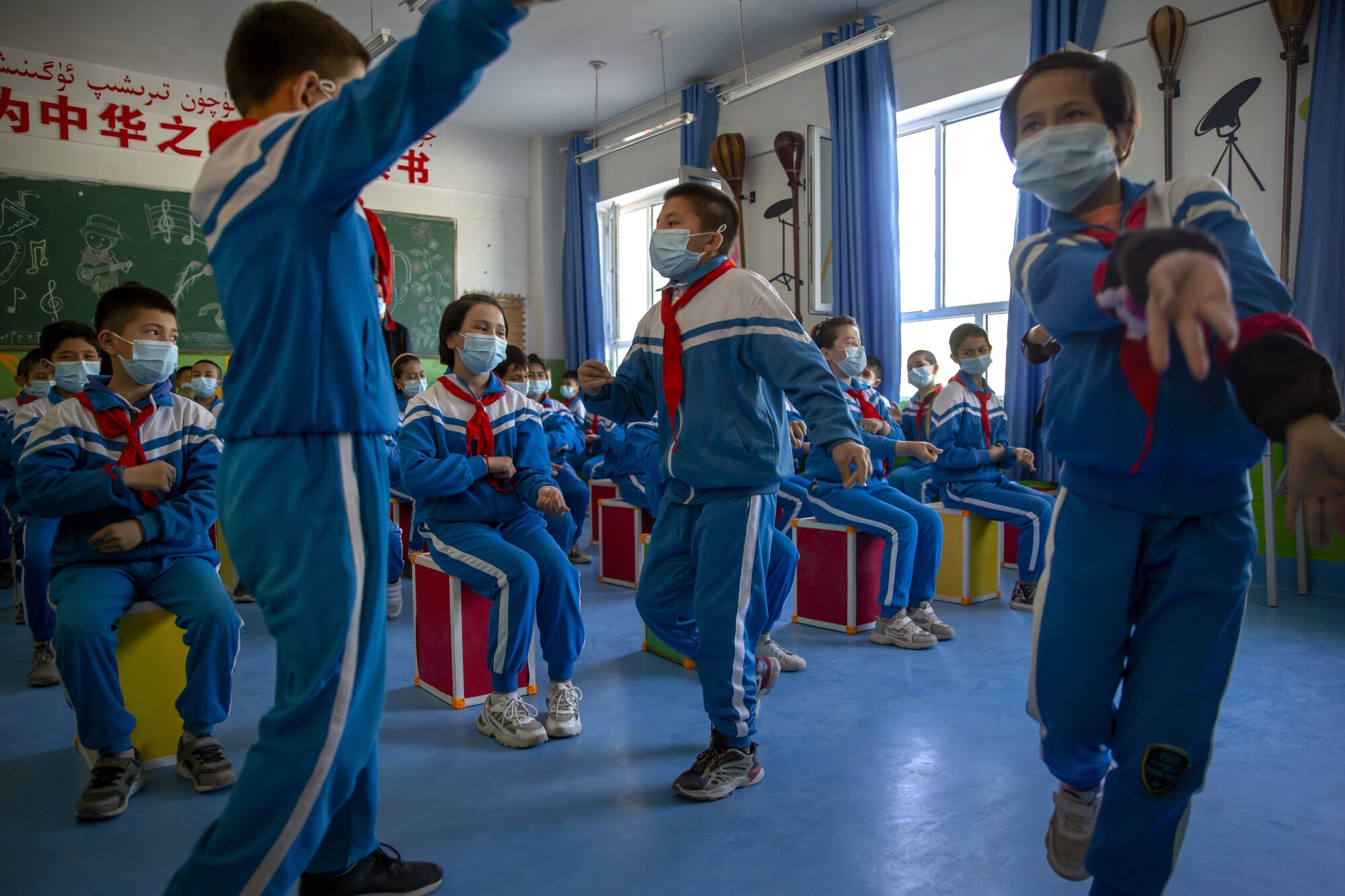 Schoolchildren in light blue and white uniforms with red scarves dance in a classroom 