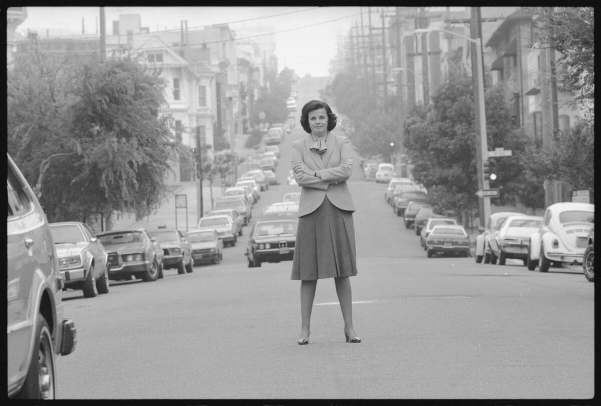 A black-and-white photo of Dianne Feinstein standing in the middle of a street with her arms crossed