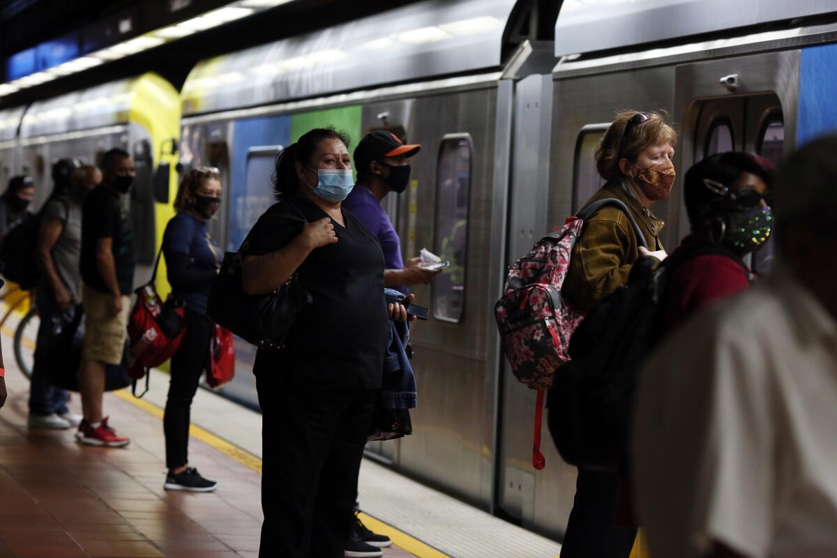 Transit riders wearing face masks and carrying backpacks stand on a train platform 