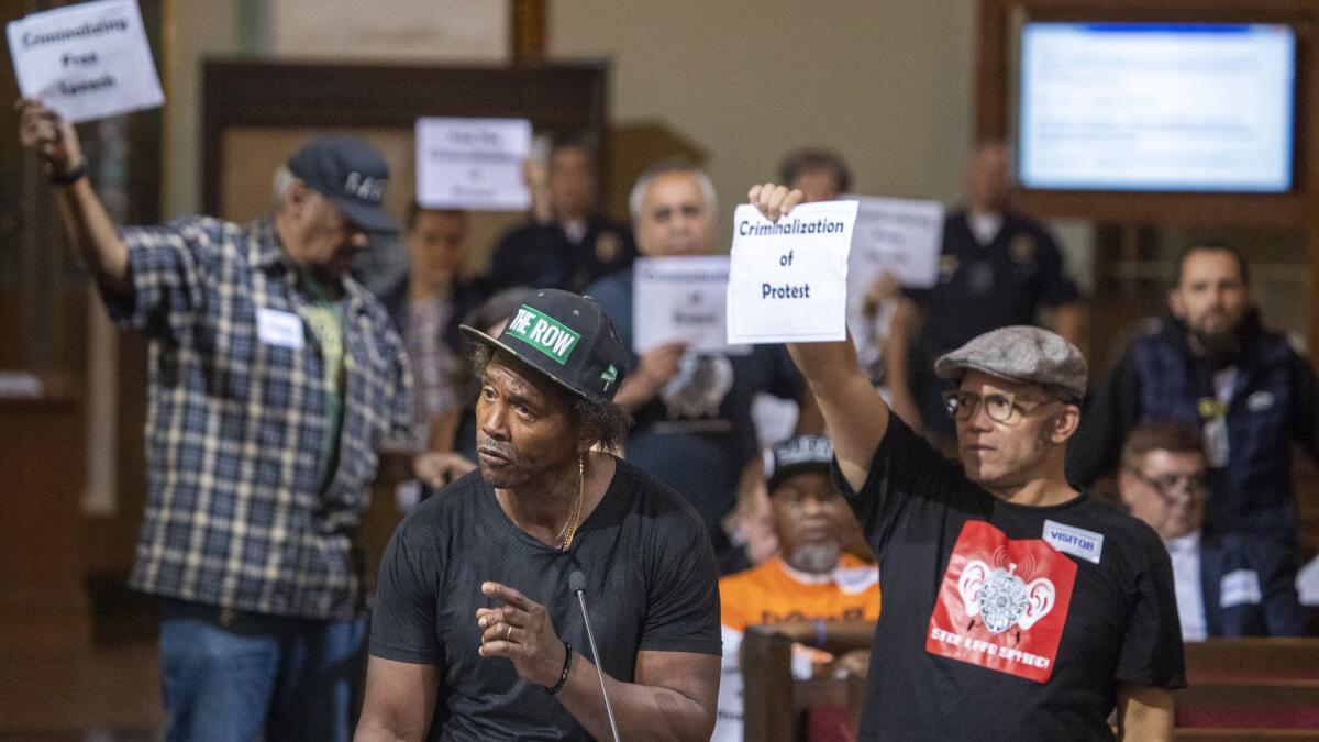 Protesters hold signs as General Dogon speaks before the Los Angeles City Council votes to crack down on people who repeatedly disrupt their meetings.