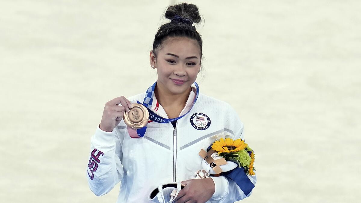 Suni Lee holds up her bronze medal and smiles.