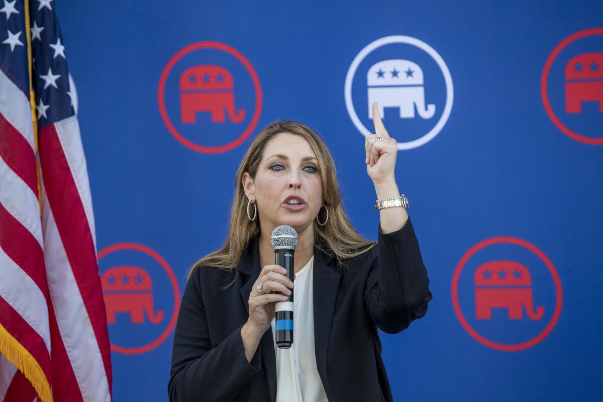 Ronna McDaniel, standing by an American flag against a blue background with stylized elephant logos, points up as she speaks