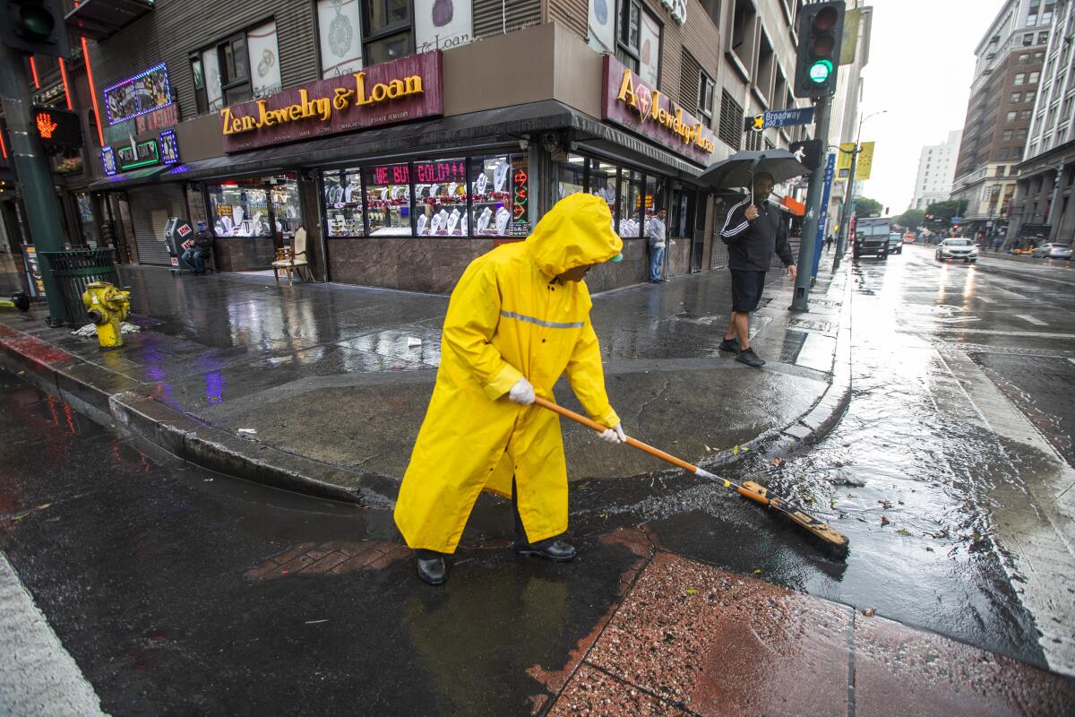 A man in a yellow raincoat clears gutters with a broom on a city corner.