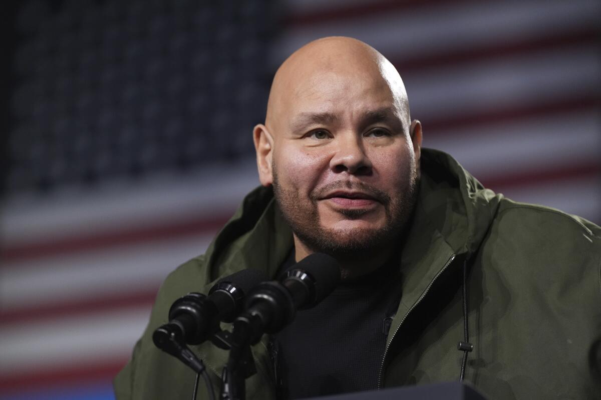 Fat Joe speaks into microphones in front of a massive American flag
