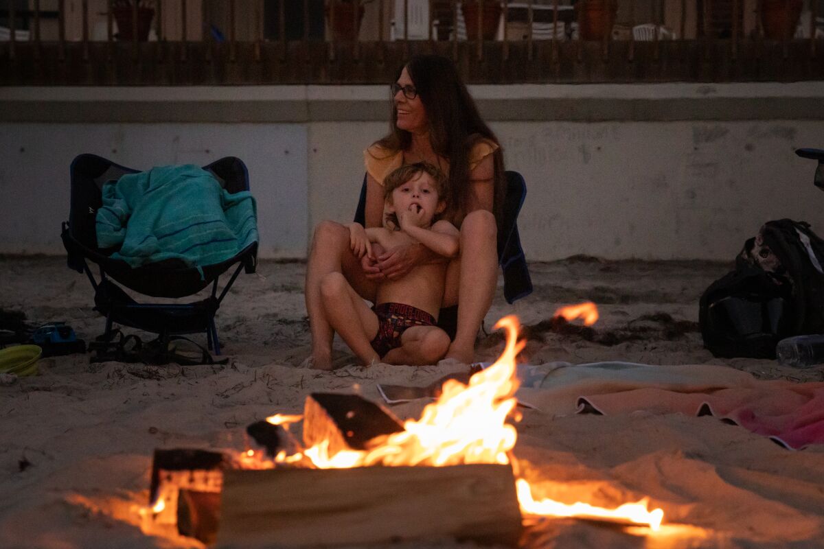 A woman and child sit on the beach in front of a small wood bonfire.