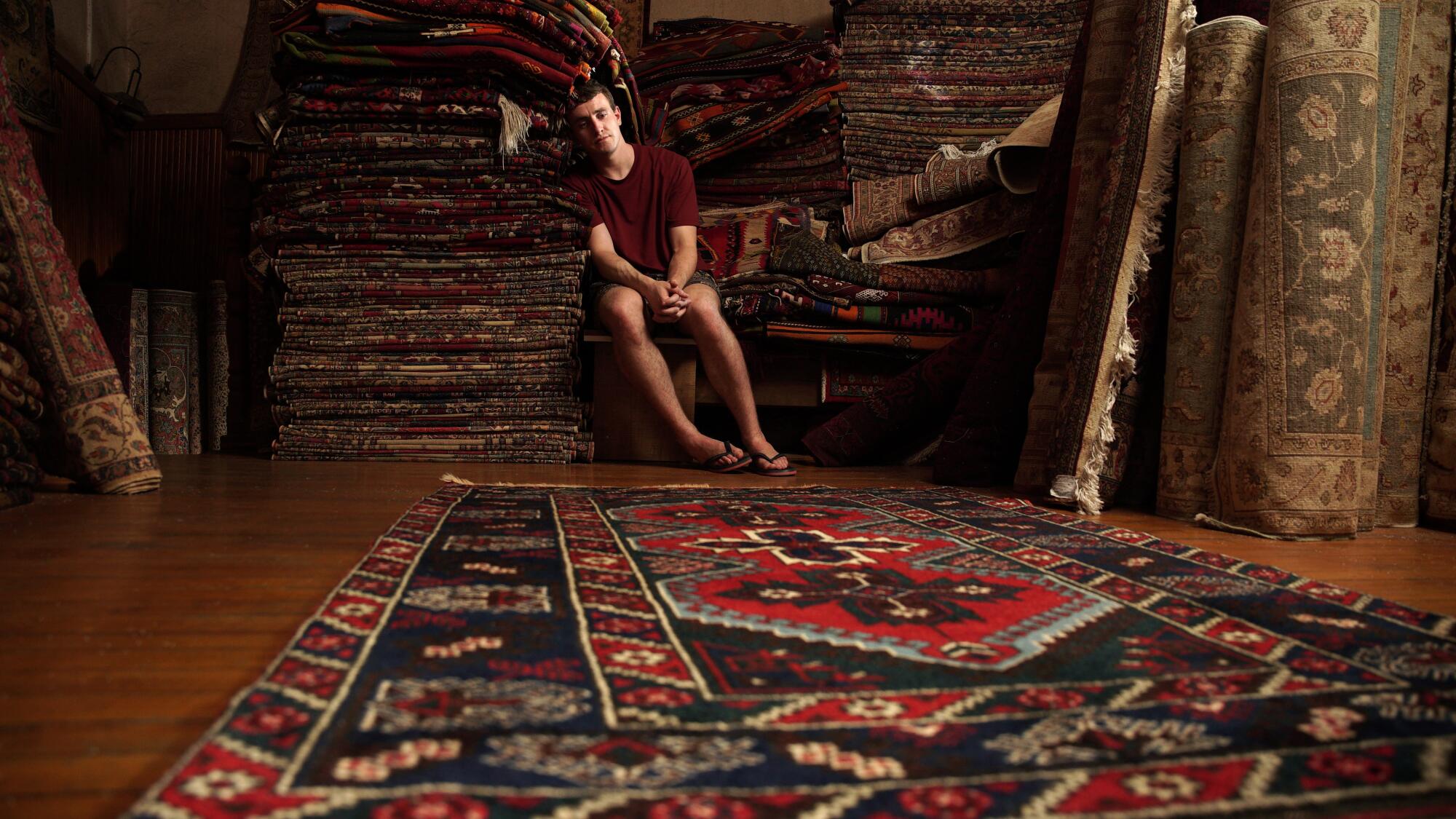 A man sits in a shop surrounded by rugs.