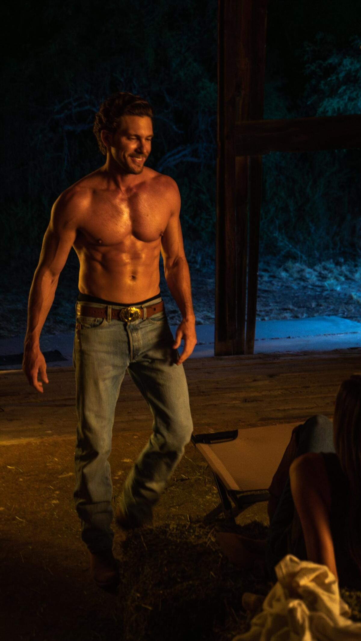 A shirtless muscular man in jeans stands outside. 