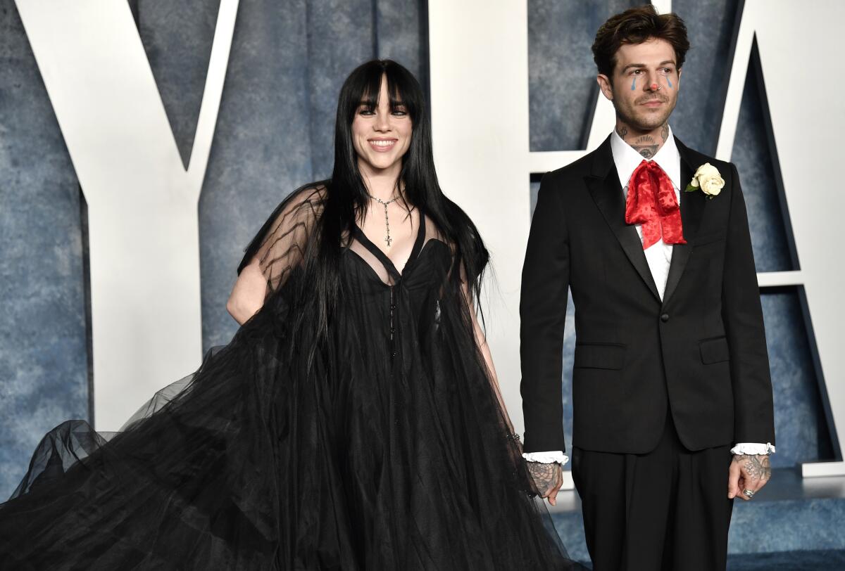 Billie Eilish in a flowing black gown, with dark hair, stands next to Jesse Rutherford in a black suit and a red ribbon tie.