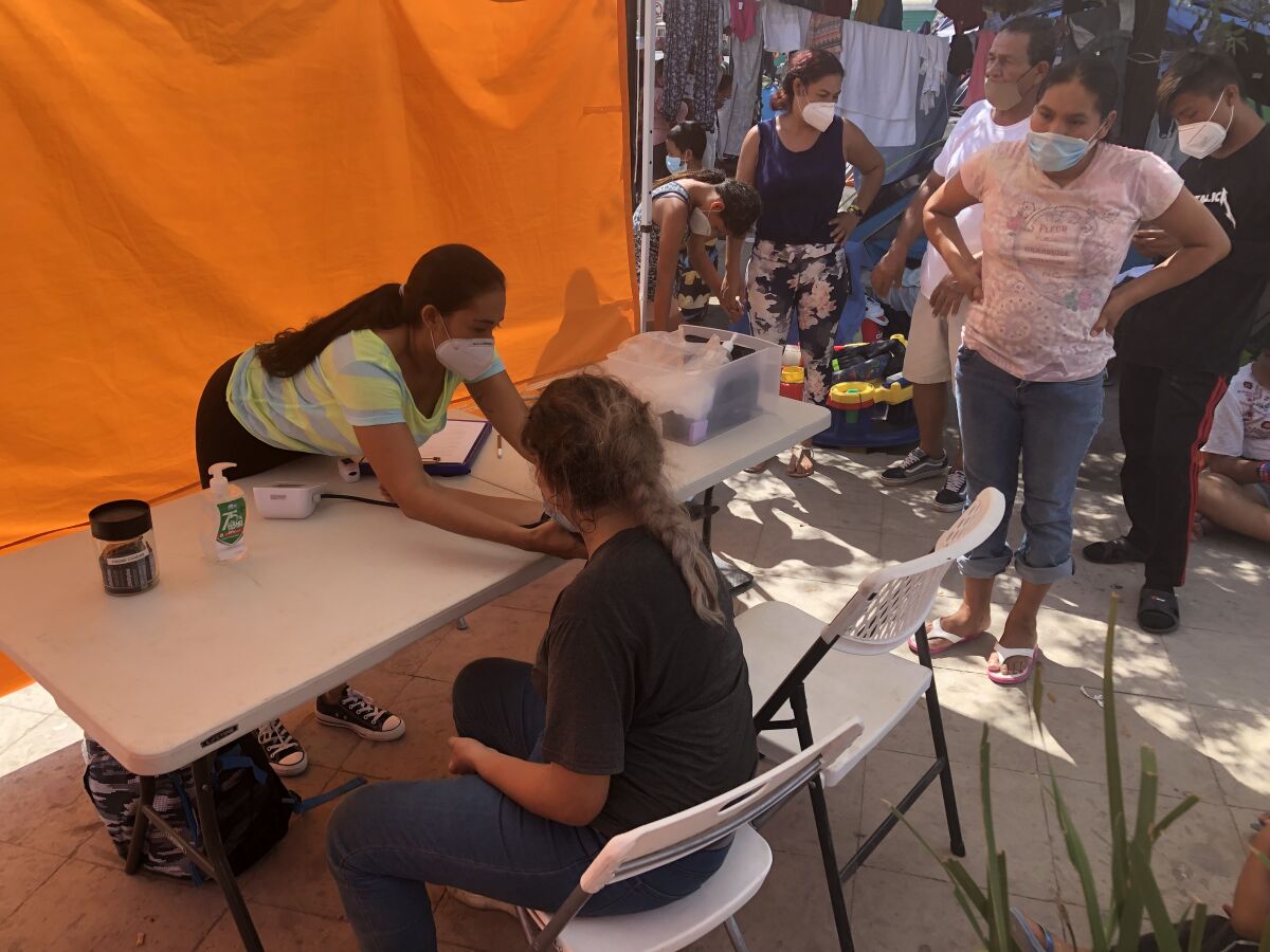A woman reaches toward a woman seated across a table at the migrant camp in Reynosa, Mexico, as others wait.