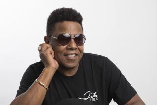 Tito Jackson in a black t-shirt and aviator sunglasses poses with his elbow on a table 