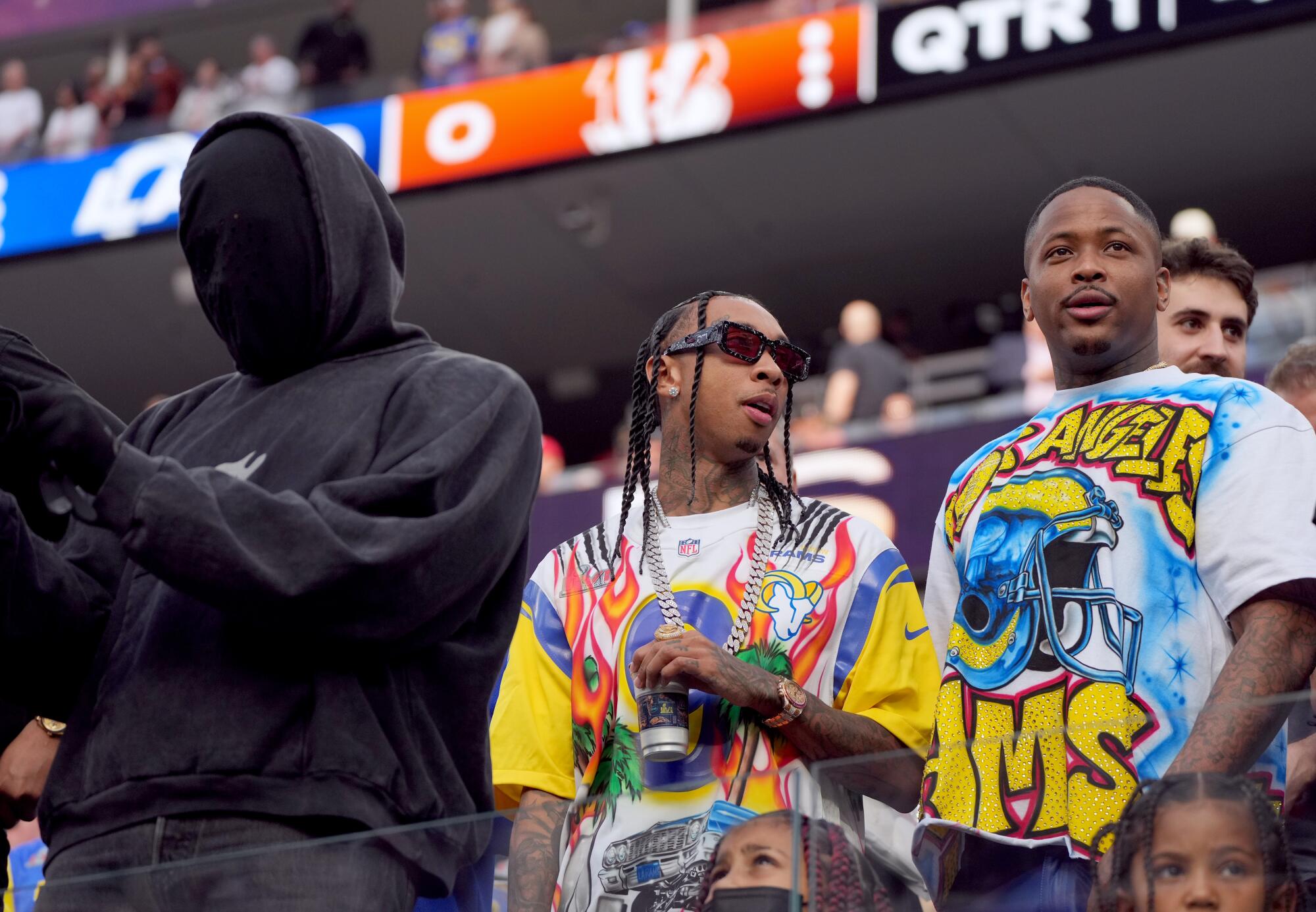A hooded man dressed entirely in black next to two men in colorful shirts at a football game.