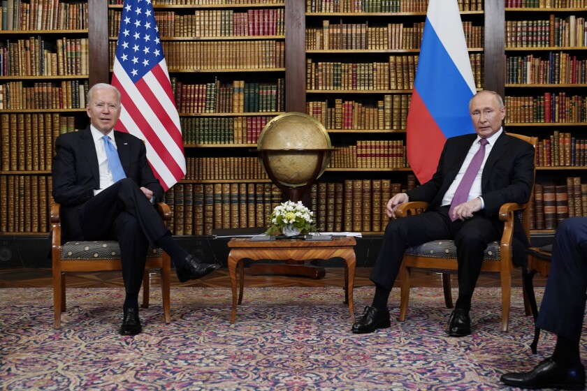 Biden and Putin sit in chairs. Between them is a coffee table and behind are U.S. and Russian flags.