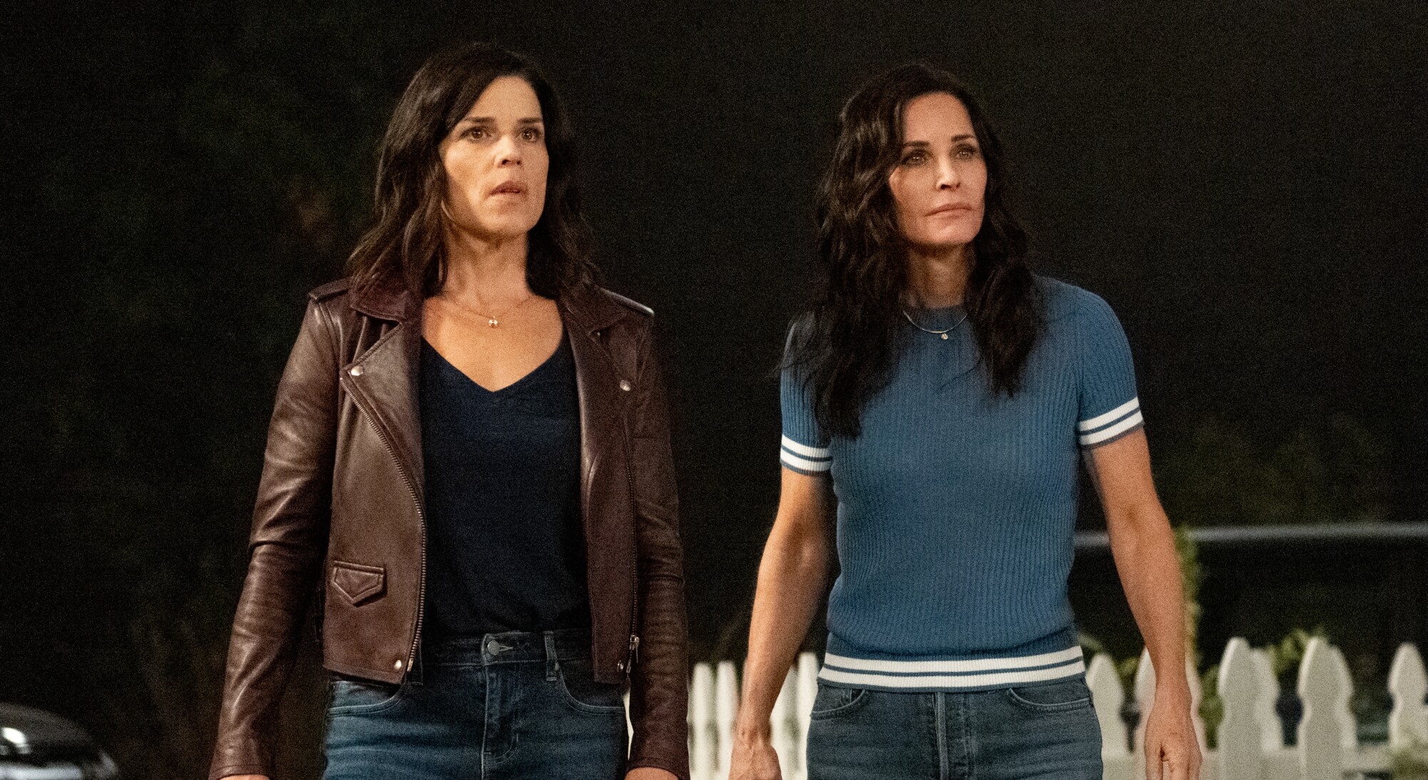 Two women stand by a white picket fence at night.