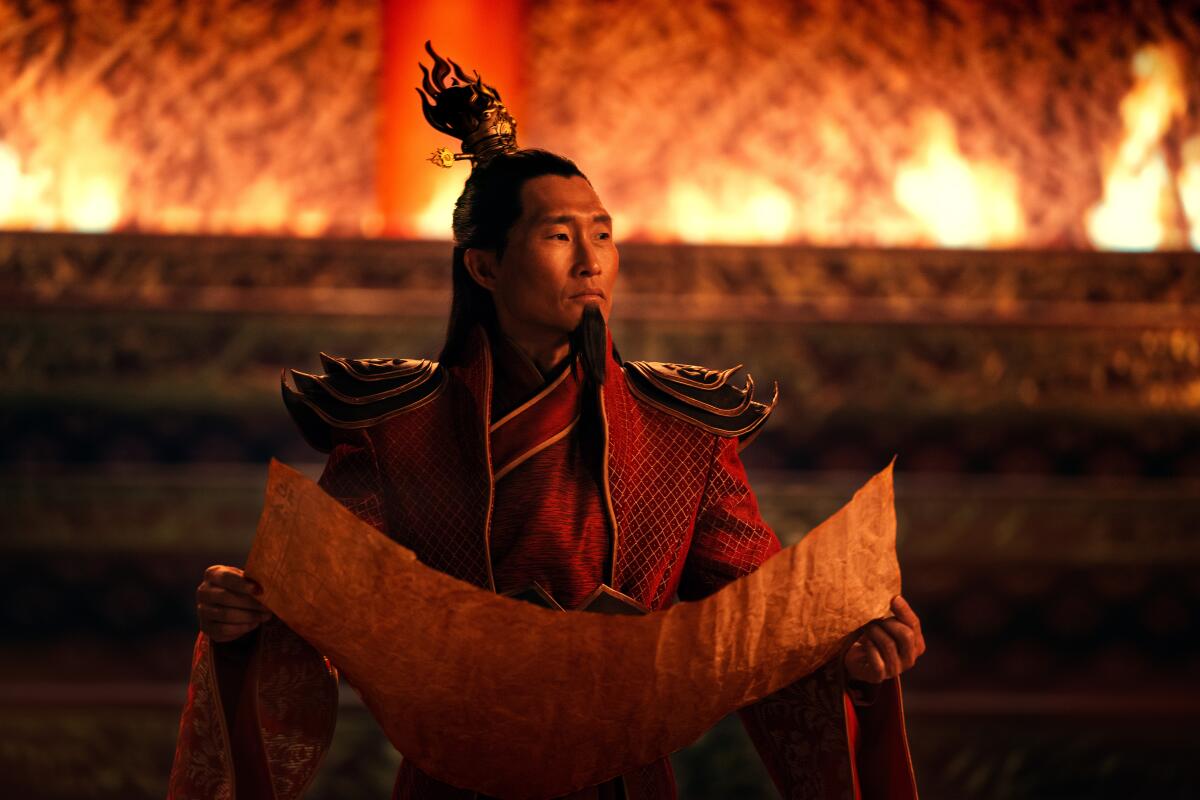A man in elaborate red costume with big epaulets stands before a wall of flame holding a scroll open before him.