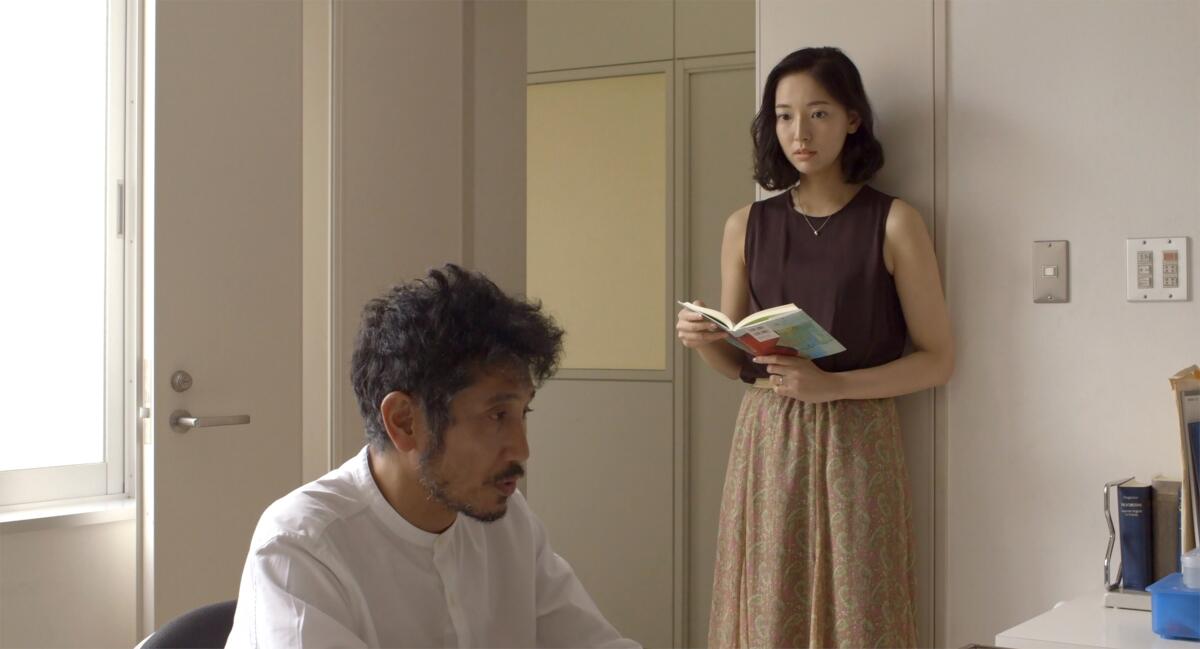 A woman holds a book while standing and facing a man seated in front of a desk