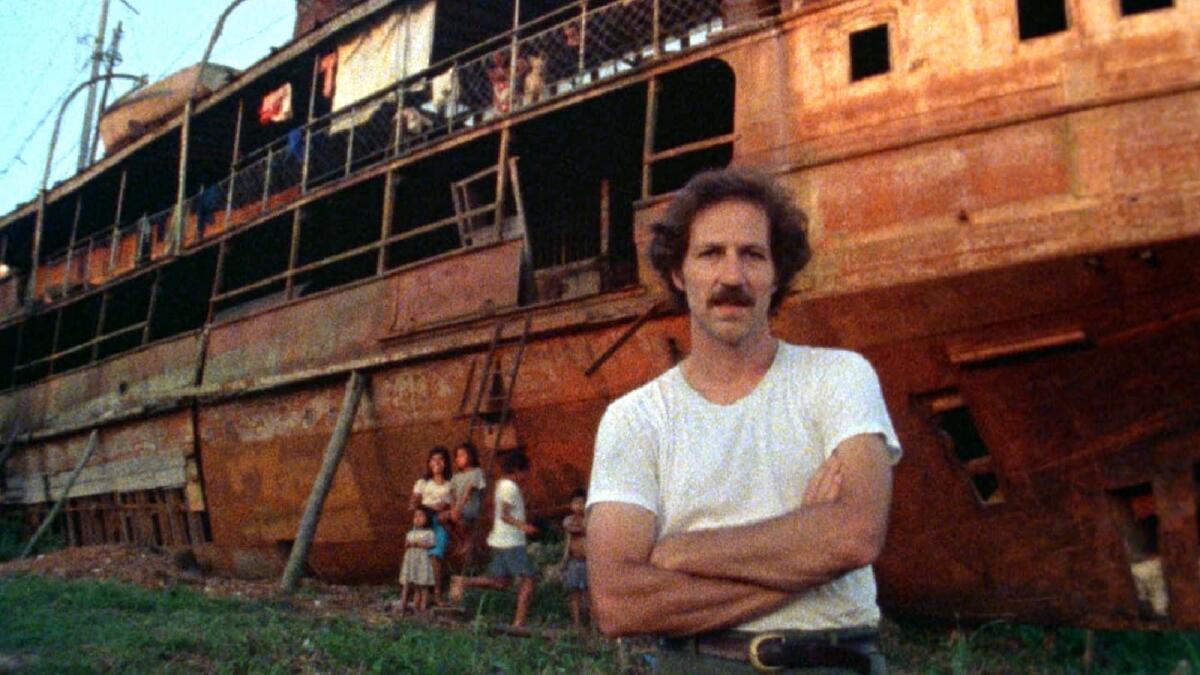 A man stands, arms crossed, in front of a large wooden boat 