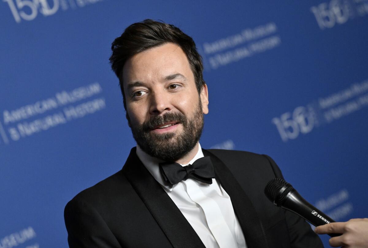 A bearded Jimmy Fallon wears a black tux and speaks into a microphone