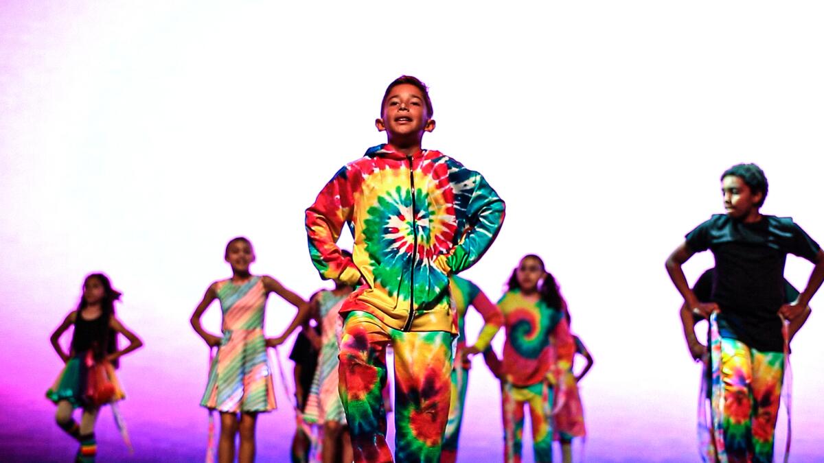 Children in tie-dyed outfits dance on a stage with purple lighting.