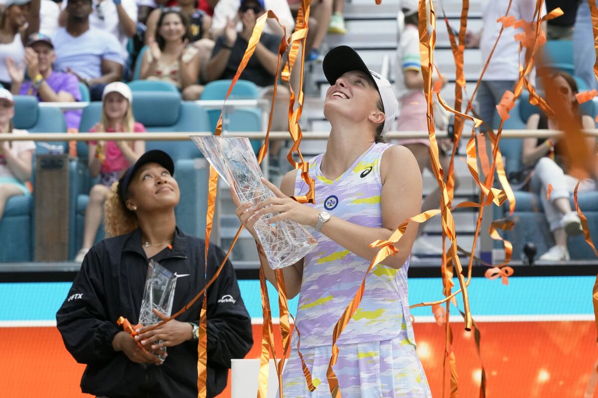 Iga Swiatek holds the winner's trophy while orange streamers fall and Naomi Osaka stands behind her.