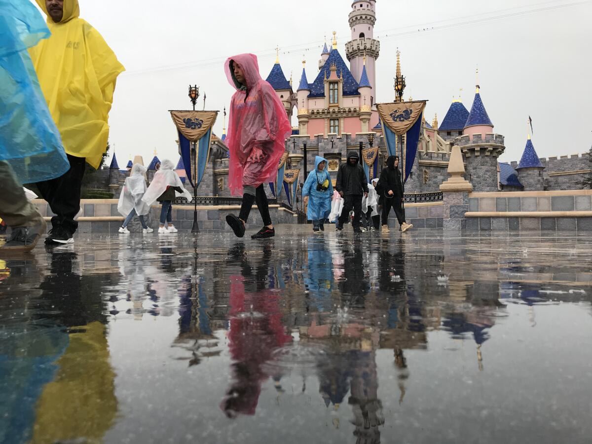 People in rain gear walk in front of Cinderella Castle amid overcast skies and puddles on the ground.