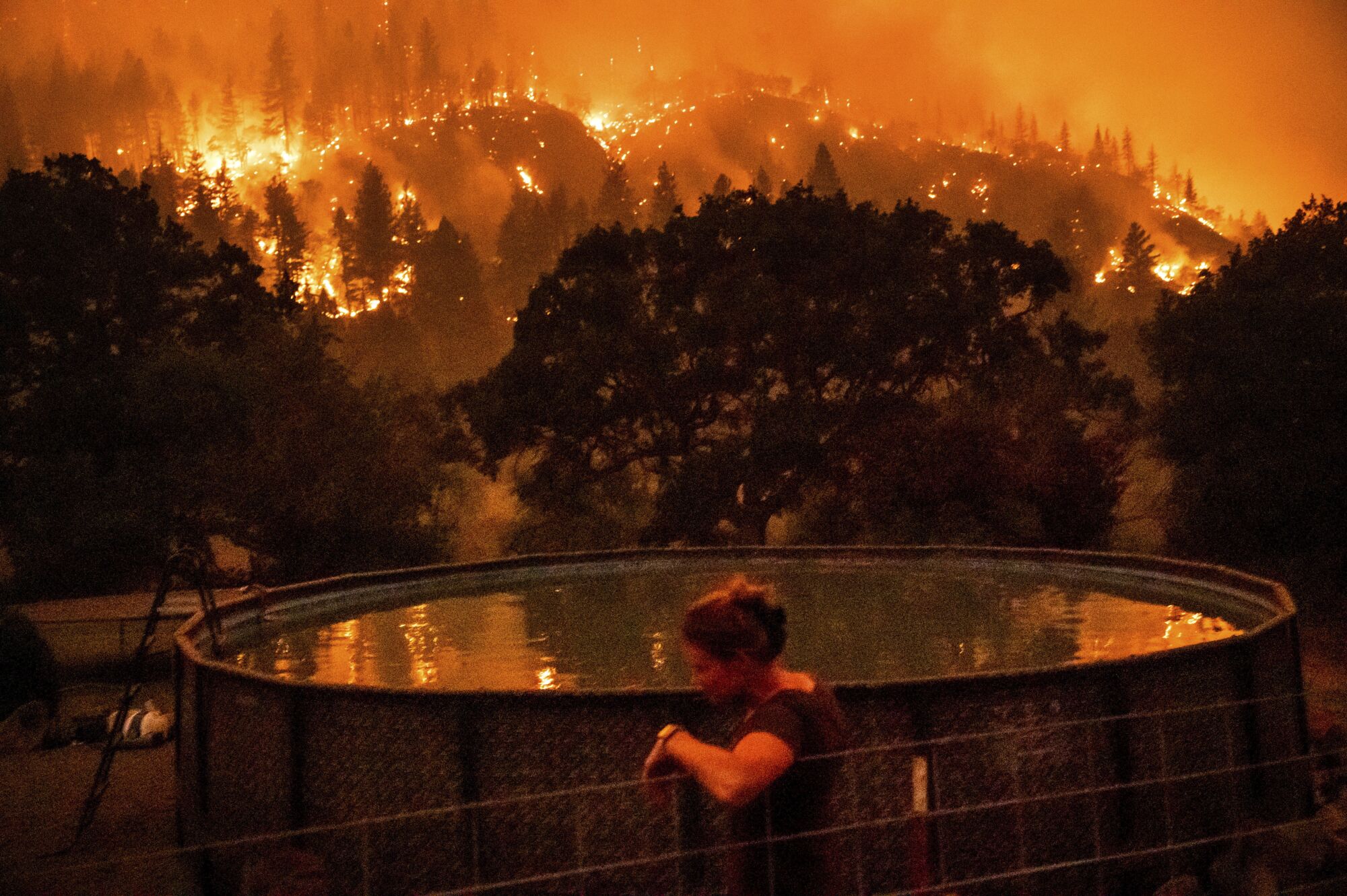 A woman leans on a fence next to an above-ground pool as wildfire burns on a nearby ridge.