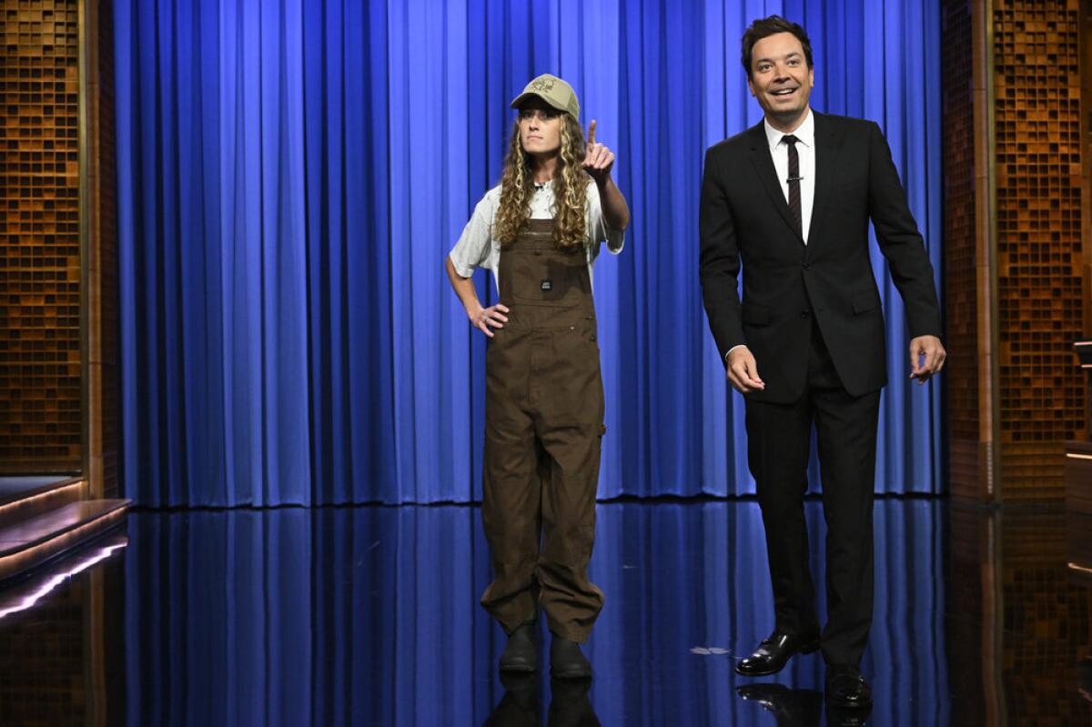 A woman wearing overalls and a hat stands beside a man wearing a suit in front of a curtain